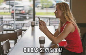 Manual de seducción: Una mujer joven y atractiva sentada sola en un café. Esta mirando su celular y tomando una bebida caliente. Tiene pelo largo, un cuerpo perfecto y un perfil de cara hermoso. 